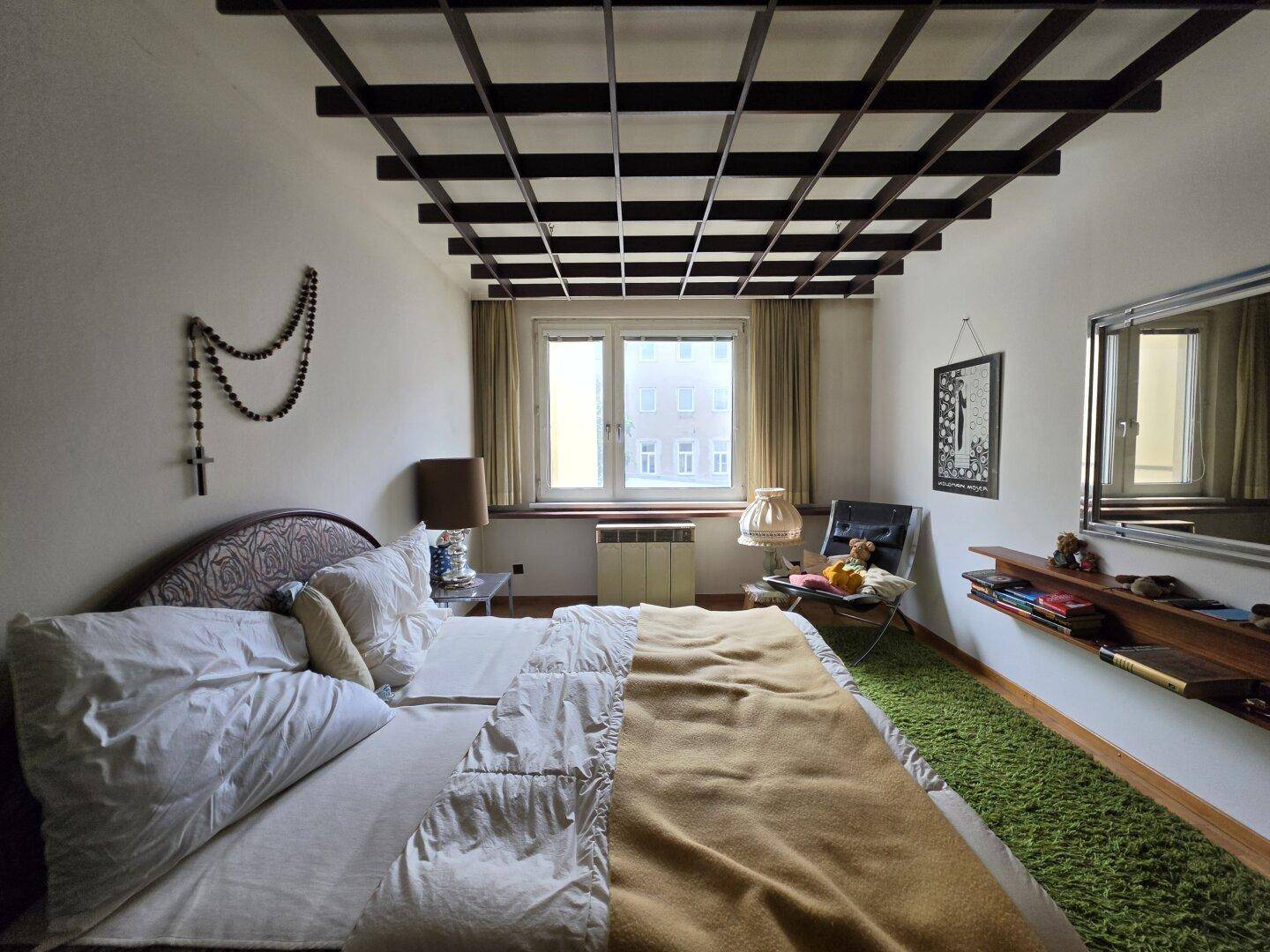 Schlafzimmer mit Gitterdecke, Teppichboden und einem Fenster, das Tageslicht hereinlässt.