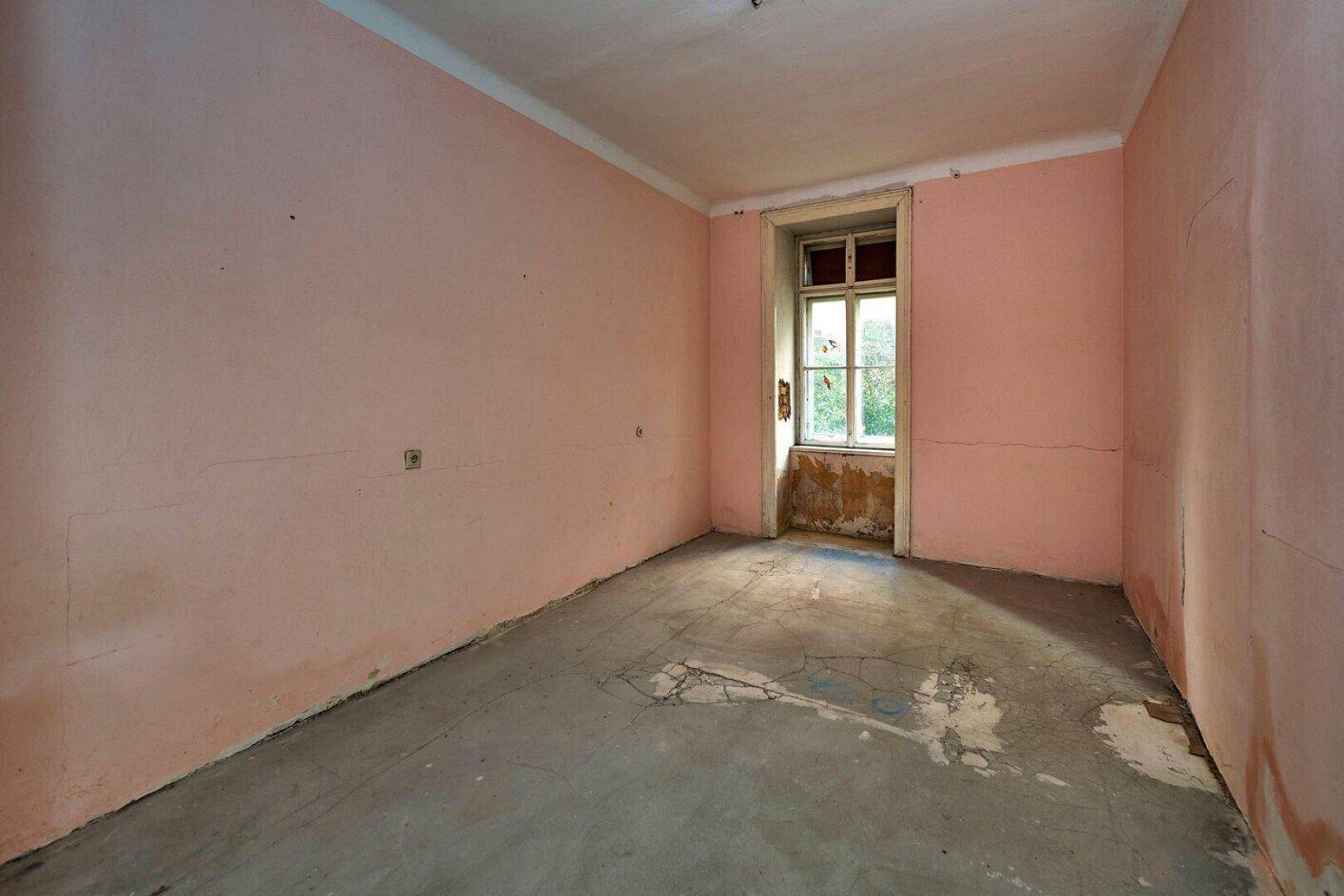 Leerer Raum mit rosa Wänden, abgenutztem Boden und einem Fenster mit Blick ins Grüne.