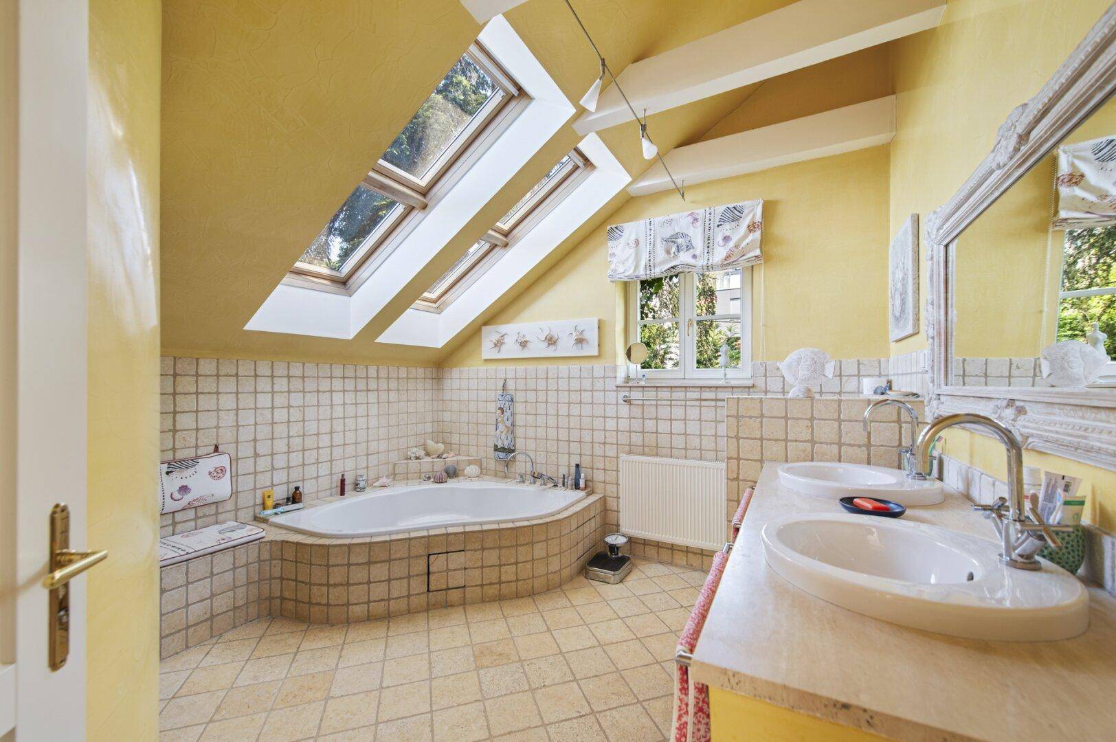 Helles Badezimmer im Dachgeschoss mit Eckbadewanne, Doppelwaschbecken, Fliesen und Dachfenstern.
