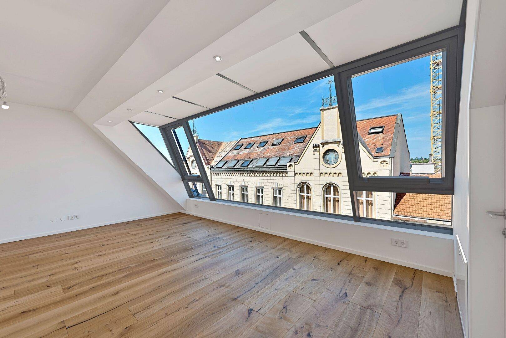 Geräumiges Zimmer mit Holzboden und breiter Fensterfront mit Blick auf die Dächer.