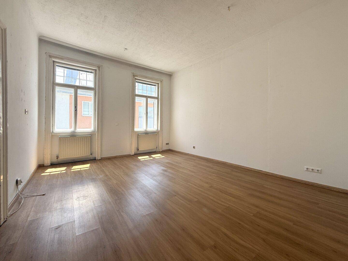 Geräumiges, leeres Zimmer mit Holzboden und zwei hohen Fenstern, die den Raum erhellen.