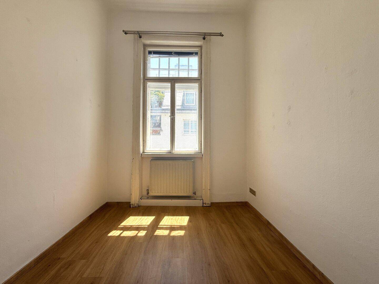 Kleines, helles Zimmer mit Holzboden und einem hohen Fenster, ideal als Schlaf- oder Arbeitszimmer.