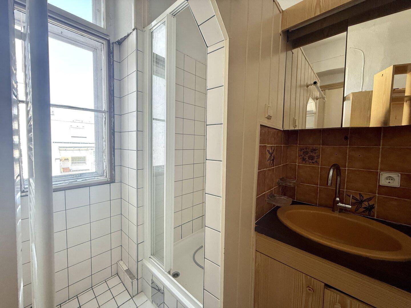 Kleines Badezimmer mit Dusche, Waschbecken und Fenster, ausgestattet mit braunen Fliesen.