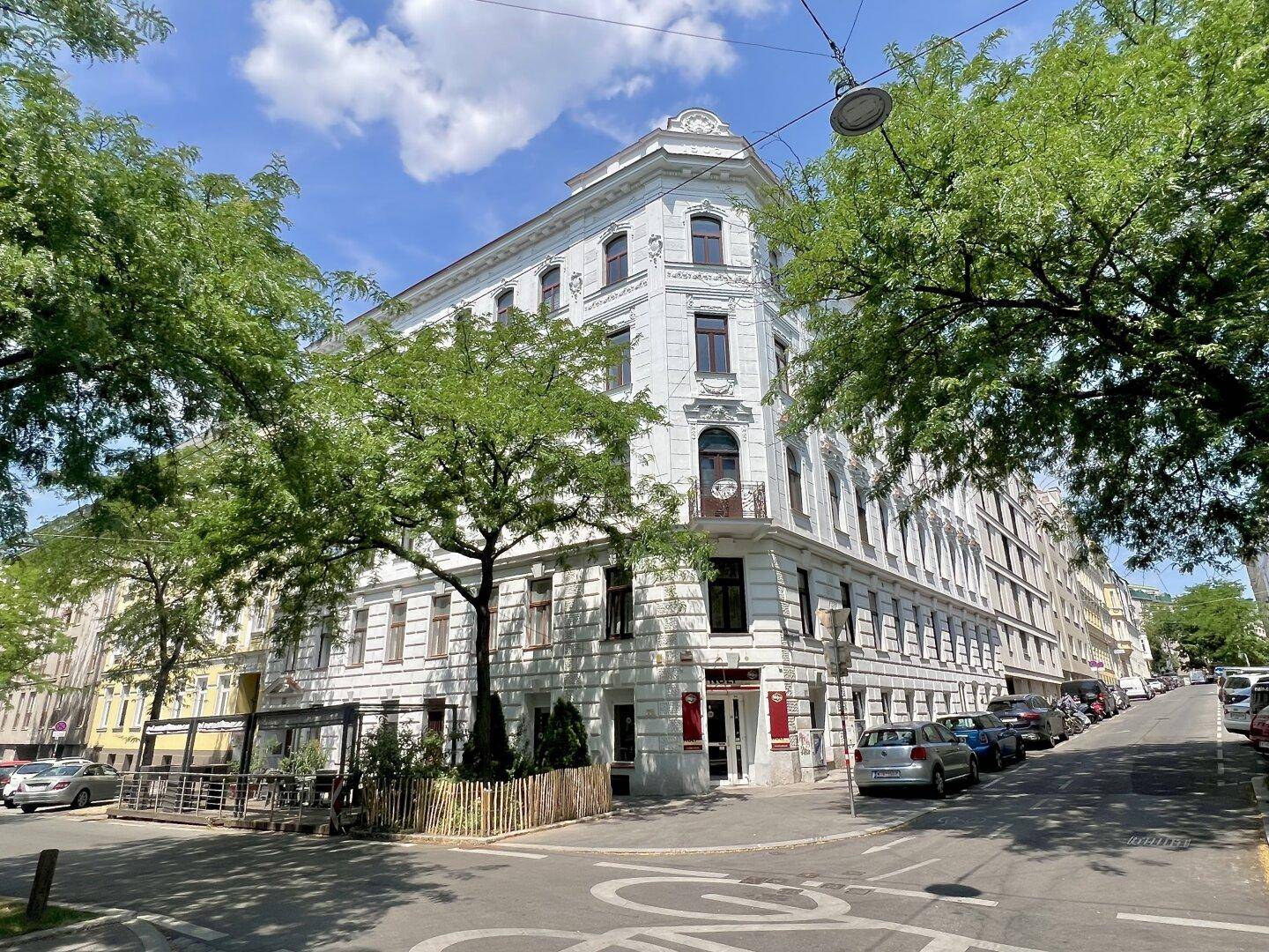 Historisches Eckgebäude mit weißer Fassade und Bäumen an einer städtischen Straße.