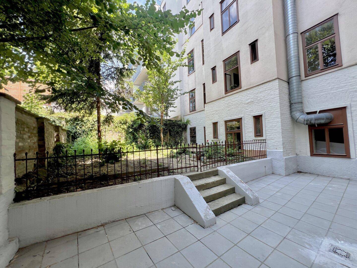 Hinterhof mit gepflasterter Terrasse und Treppe, die zu einem grünen Gartenbereich führt.