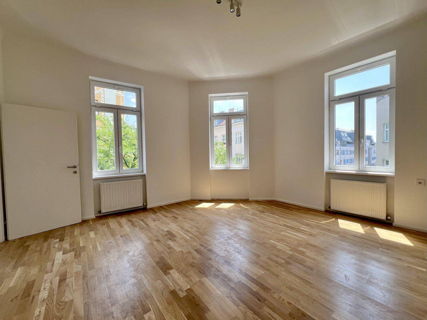 Geräumiger, heller Raum mit Holzboden, hohen Decken und großen Fenstern.