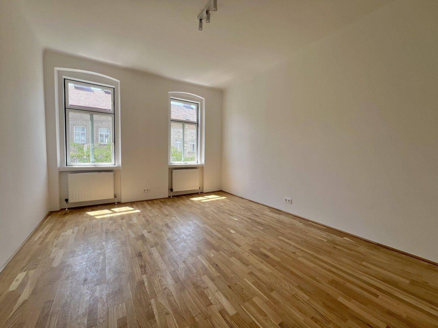 Helles, leeres Zimmer mit Holzboden und zwei Fenstern, die viel Tageslicht hereinlassen.