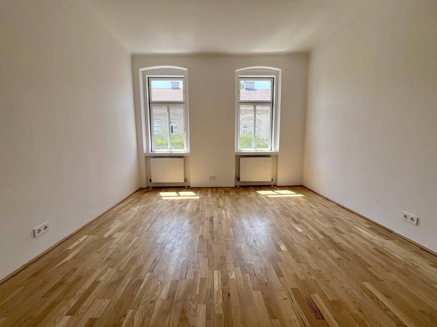 Großes, lichtdurchflutetes Zimmer mit hellem Parkettboden und zwei hohen Fenstern.