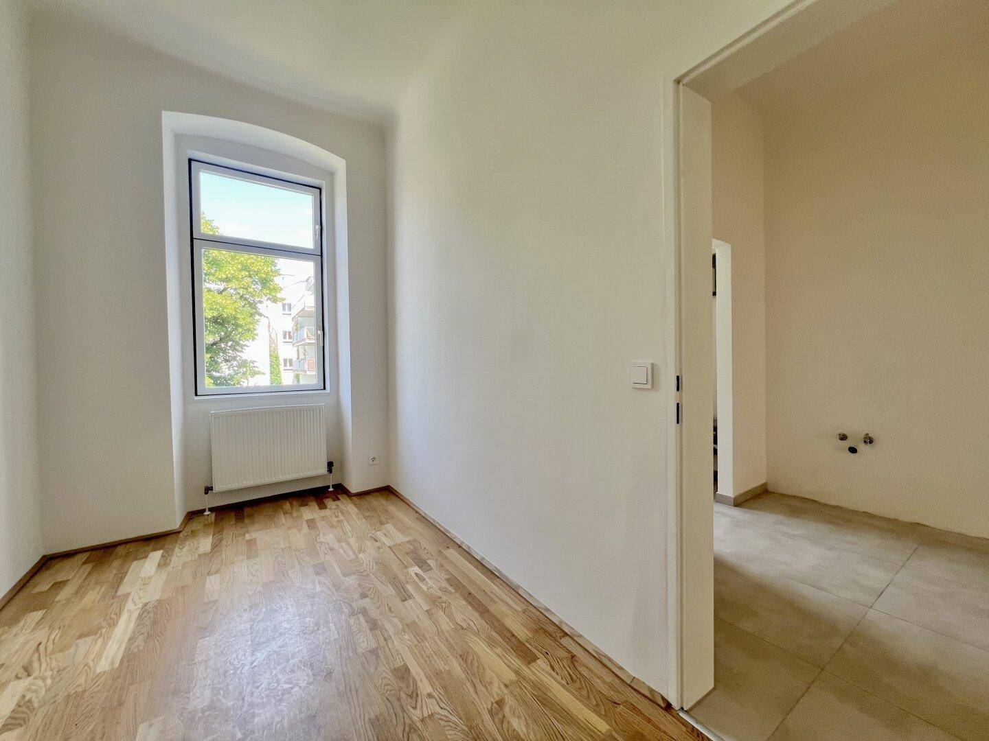 Helles Zimmer mit Holzboden und einem Fenster, das zu einem gefliesten Bereich führt.