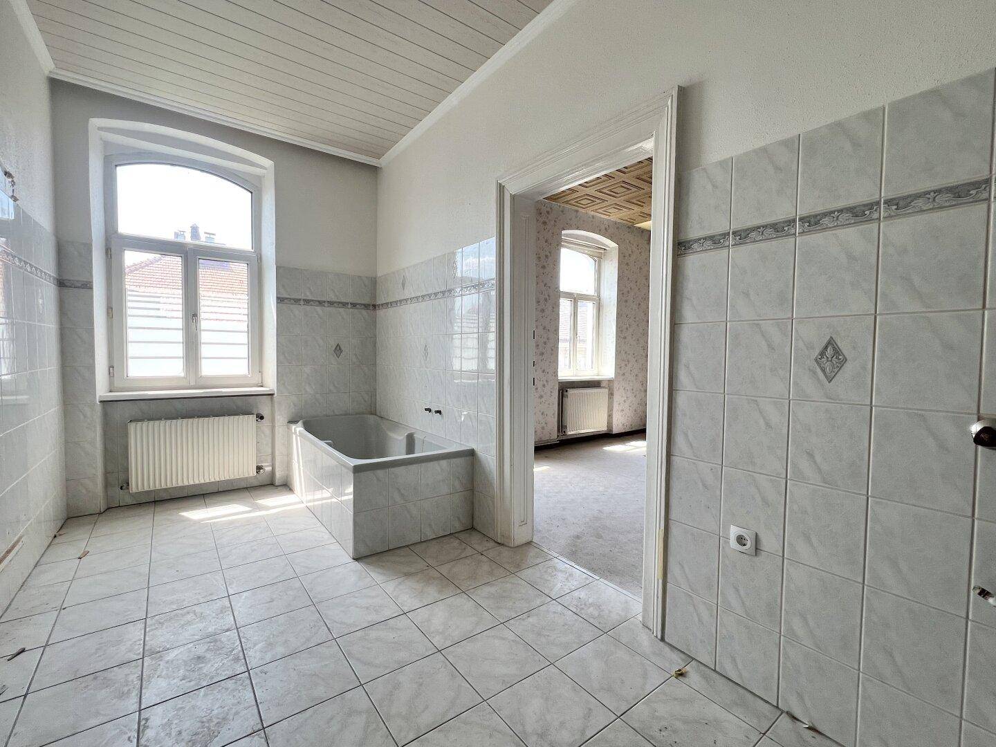 Großes Badezimmer mit Fliesenboden, eingebauter Badewanne und Fenster, bereit zur Modernisierung.
