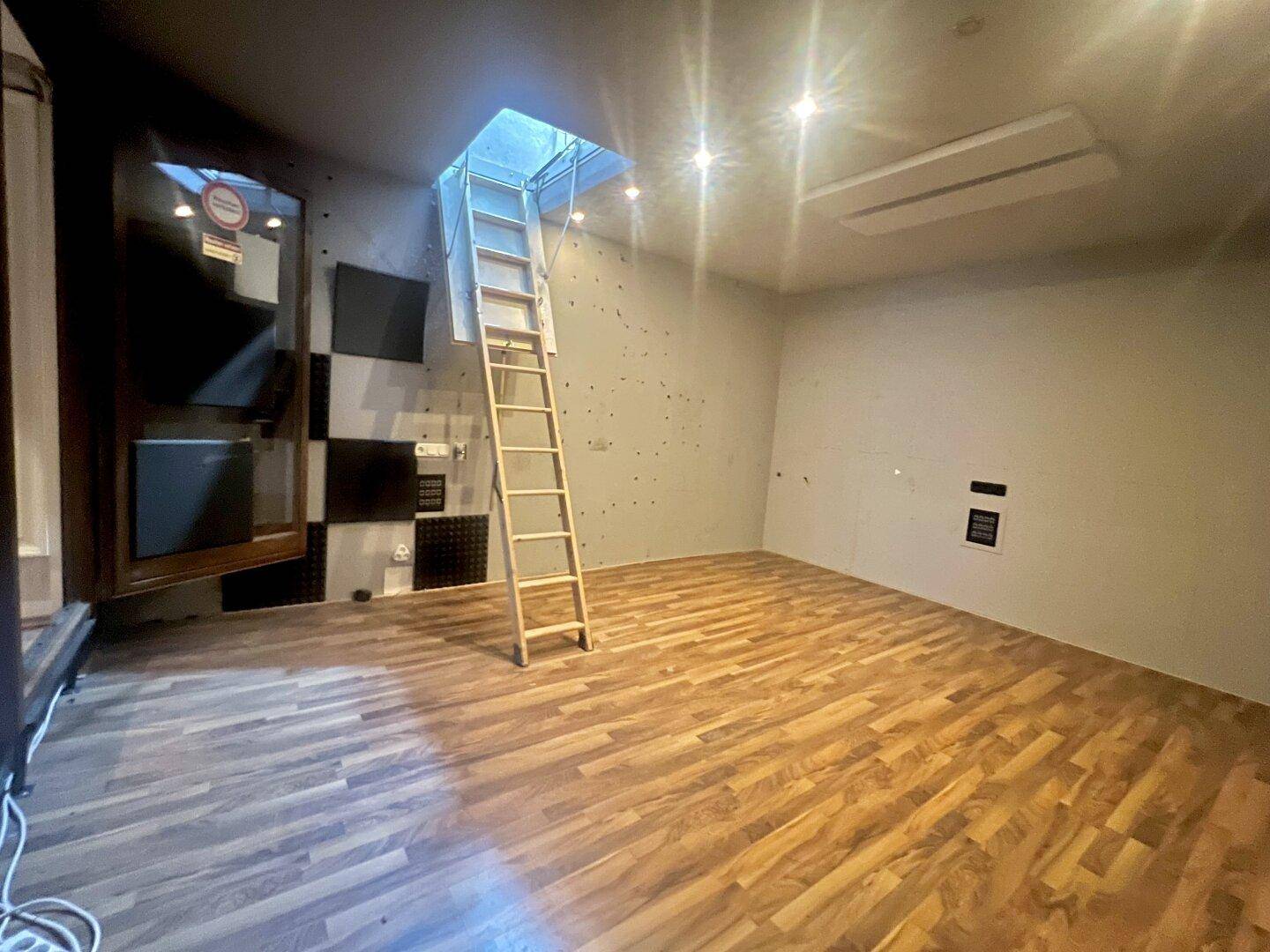 Leerer Raum mit Holzboden, einer Leiter zu einem Dachfenster und schallabsorbierenden Paneelen an der Wand.