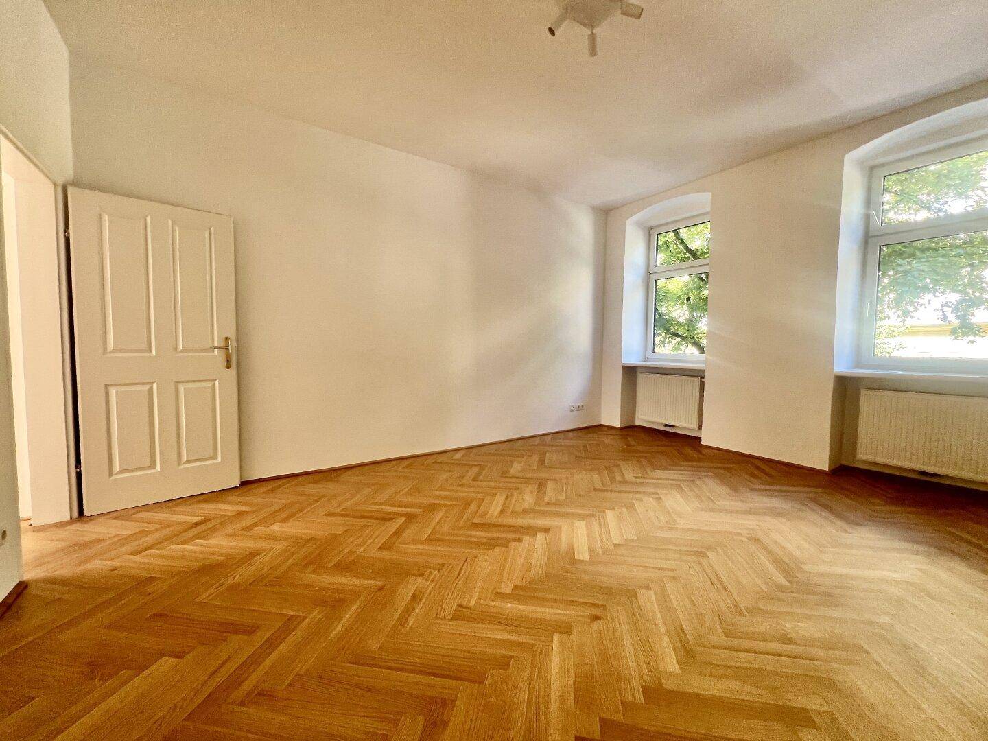 Helles Zimmer mit Fischgrätparkett, zwei Fenstern und einer weißen Holztür.