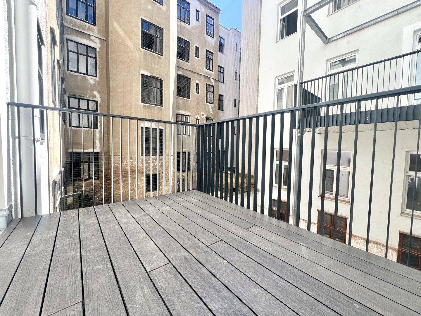Moderner Balkon mit Holzoptik-Boden und Metallgeländer, umgeben von Gebäuden.