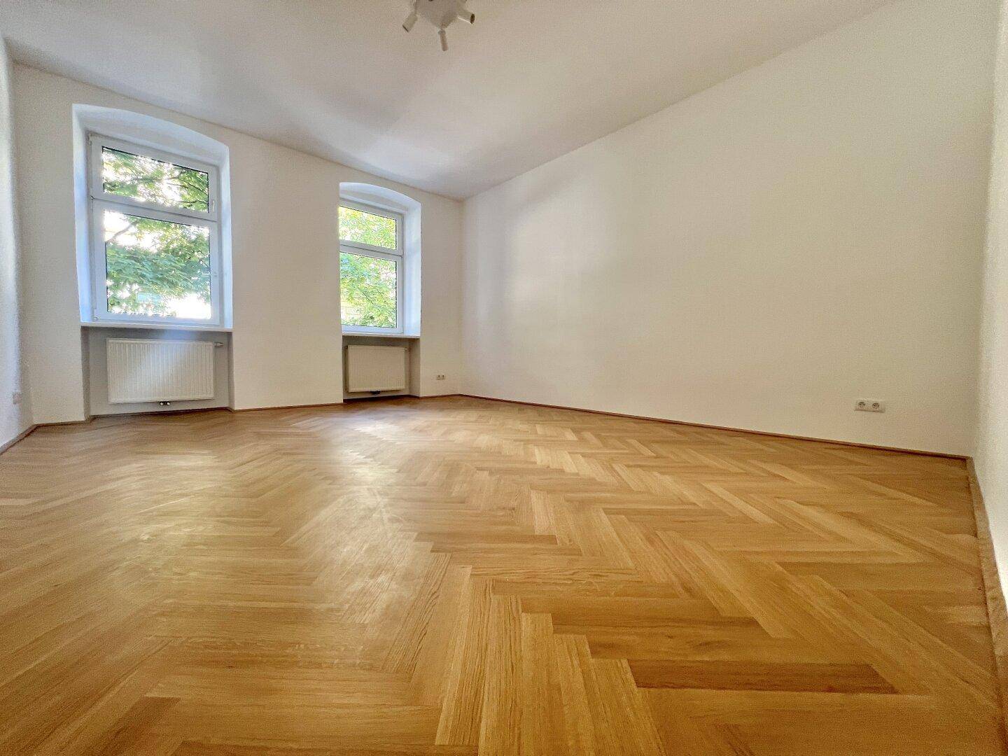 Geräumiges Zimmer mit Fischgrätparkett und zwei großen Fenstern, die den Raum erhellen.