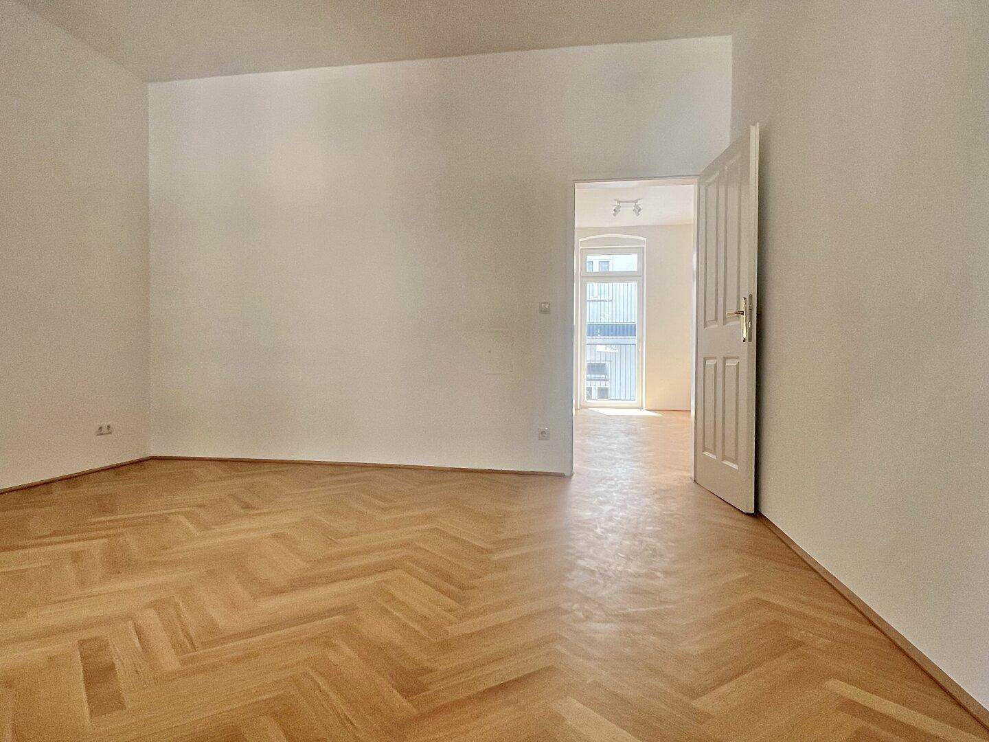 Geräumiges Zimmer mit Fischgrätparkett und Zugang zu einem weiteren hellen Raum.