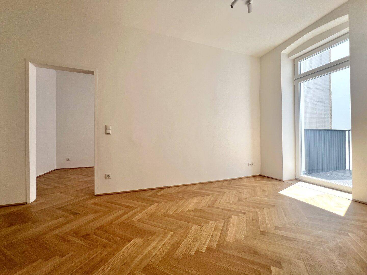 Heller Raum mit Fischgrätparkett und Zugang zu einem Balkon, der viel Licht spendet.