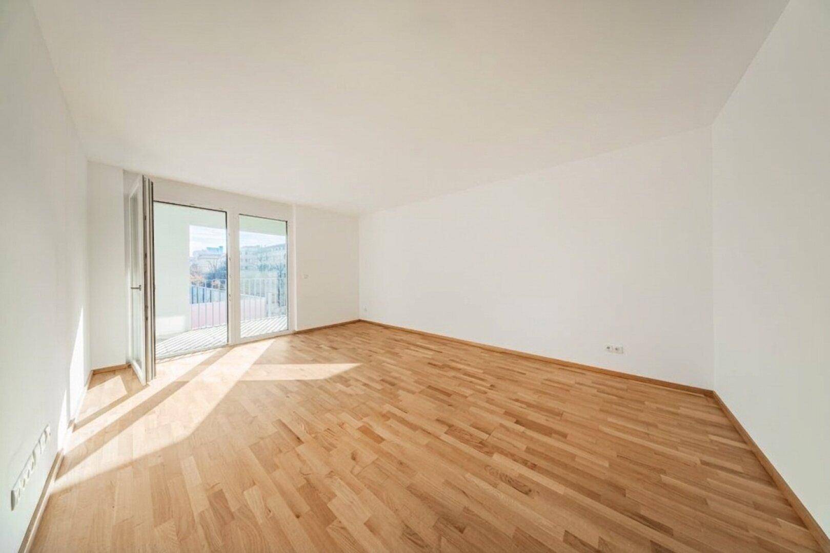 Helles, leeres Zimmer mit Holzboden und Zugang zu einem Balkon.