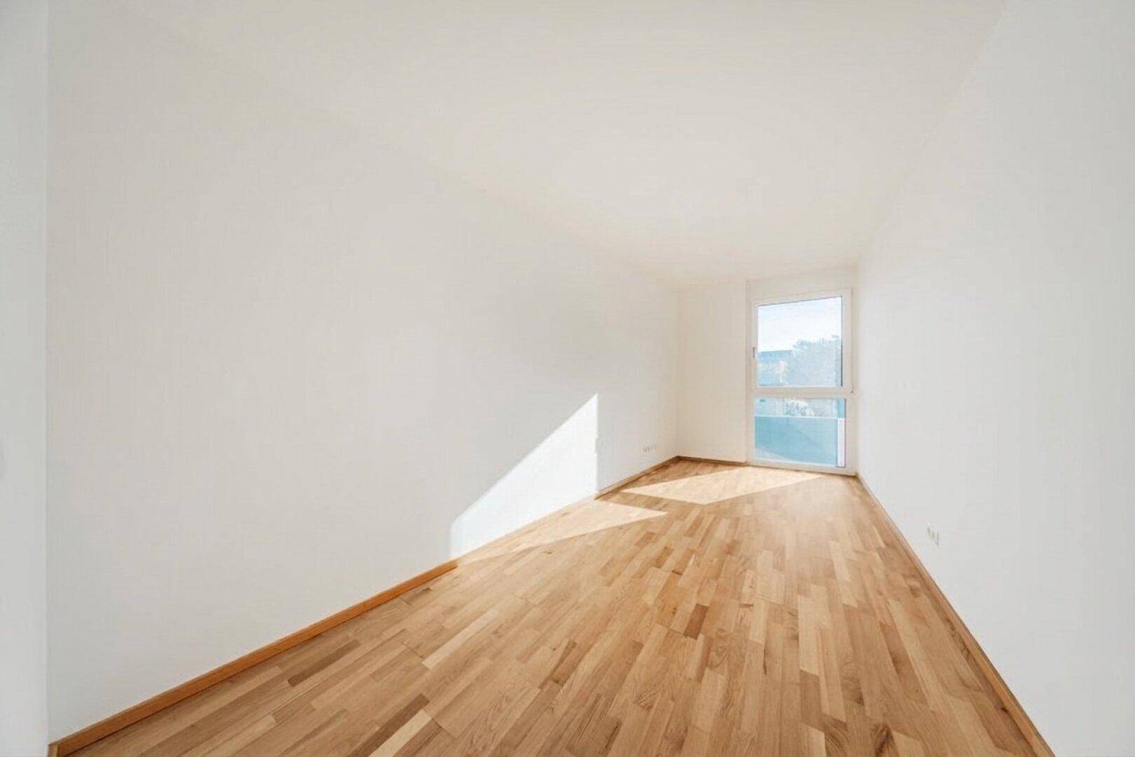 Helles, leeres Zimmer mit Holzboden und großem Fenster, ideal als Schlafzimmer.