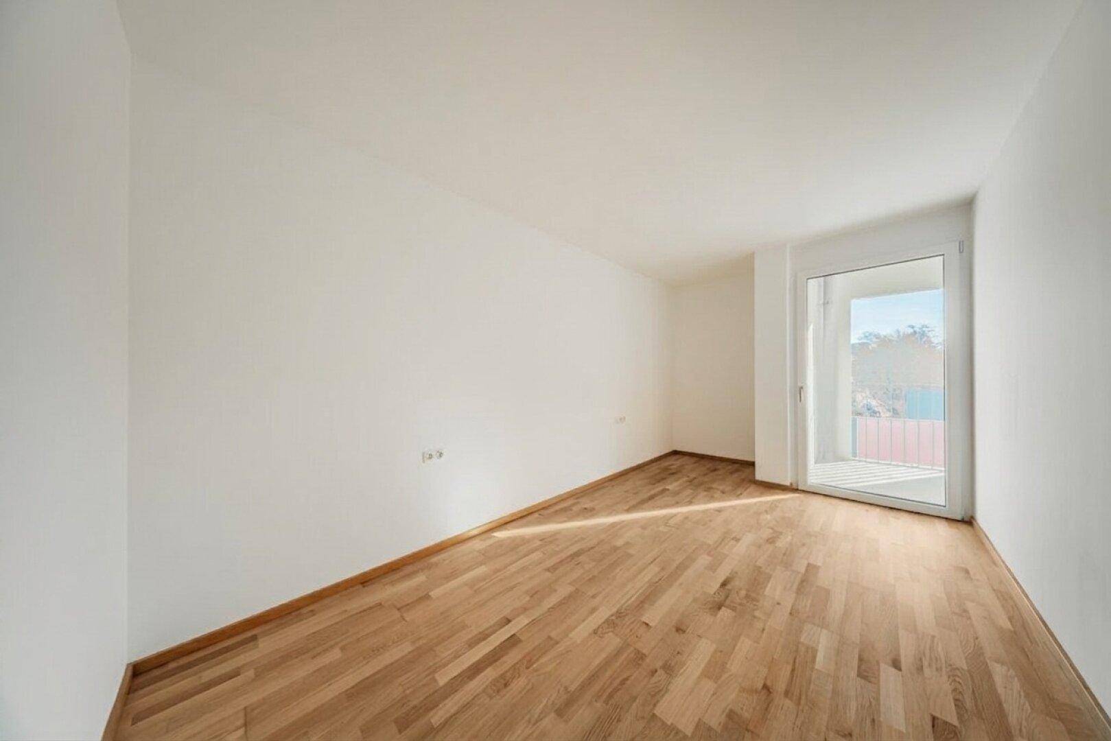 Helles, leeres Zimmer mit Holzboden und Zugang zu einem Balkon.