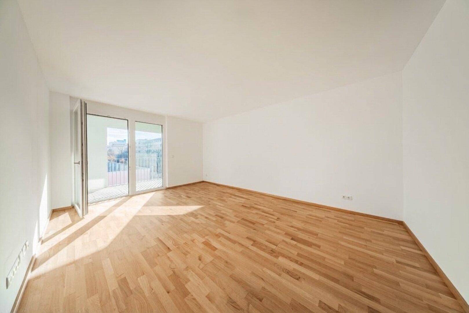 Geräumiger, leerer Raum mit hellem Holzboden und großen Fenstern zum Balkon.