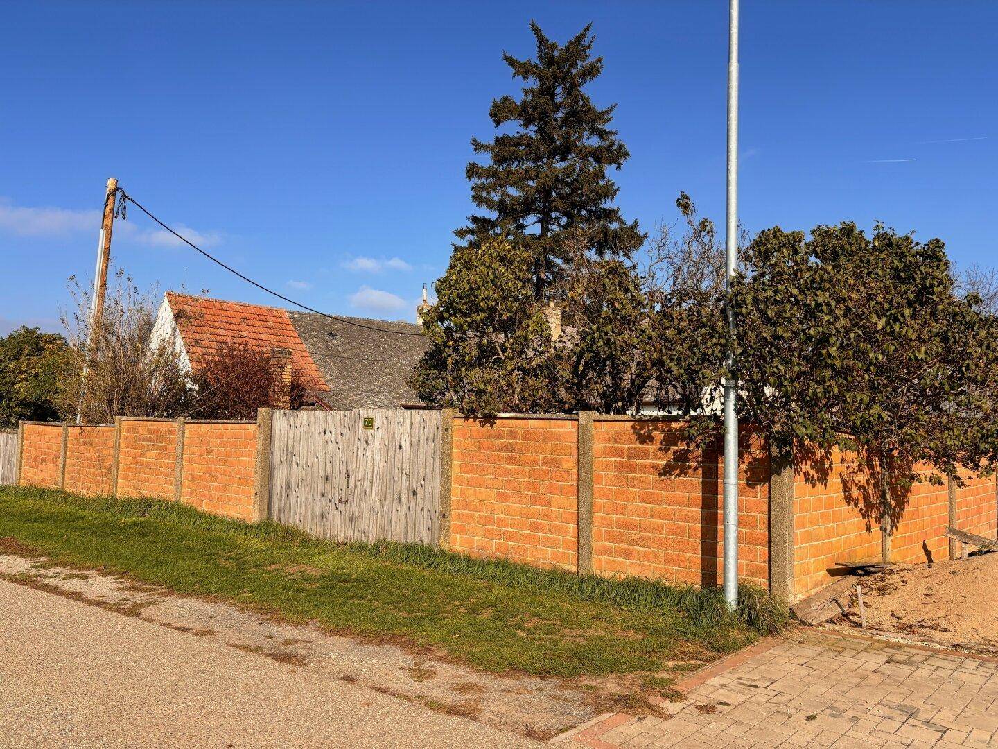 Ziegelmauer als Grundstücksbegrenzung mit Holztor und Blick auf ein Hausdach im Hintergrund.