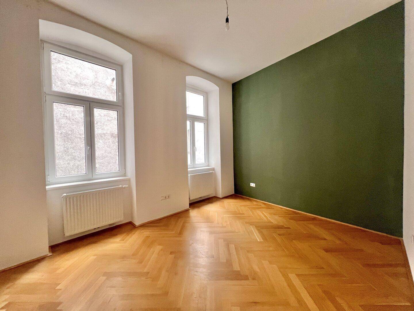 Geräumiger, heller Raum mit Fischgrätparkett, zwei Fenstern und einer auffälligen grünen Akzentwand.