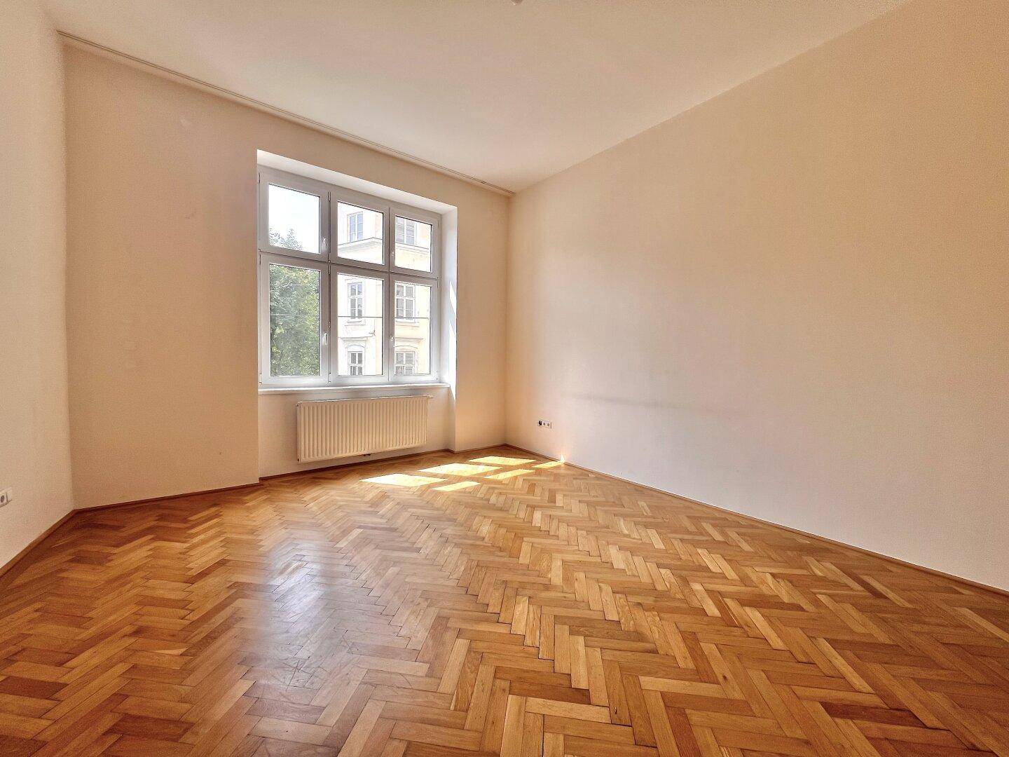 Lichtdurchfluteter Raum mit klassischem Fischgrätparkett und einem großen Fenster, das den Raum erhellt.
