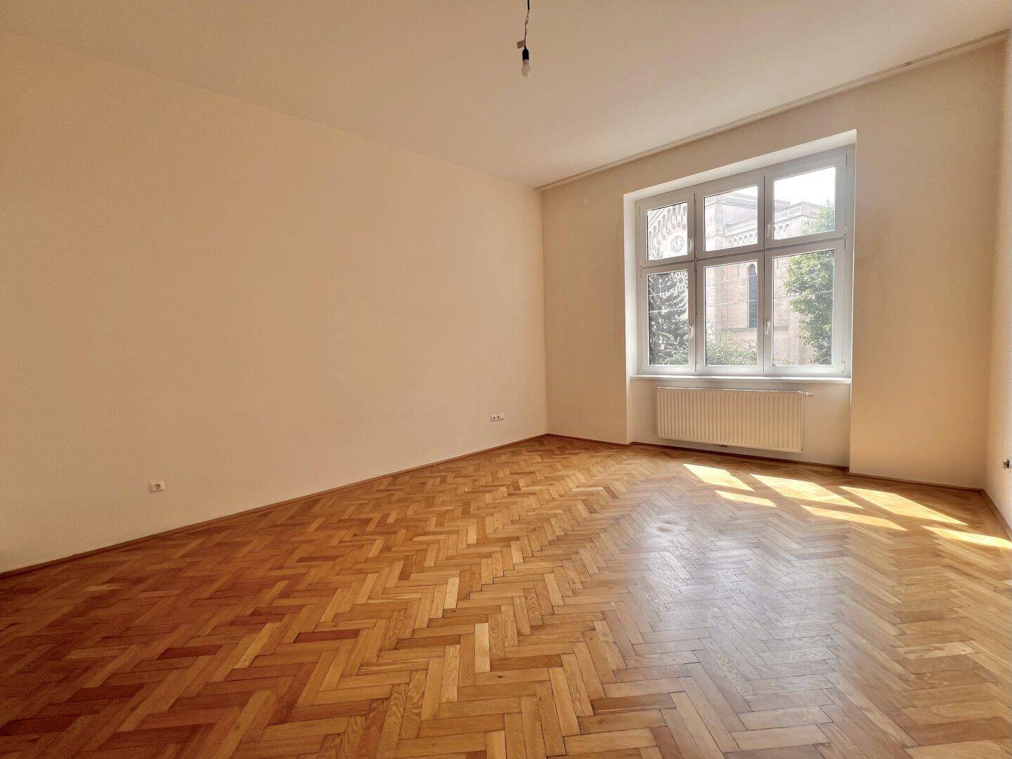 Heller, leerer Raum mit Fischgrätparkettboden und großem Fenster, das viel Tageslicht hereinlässt.