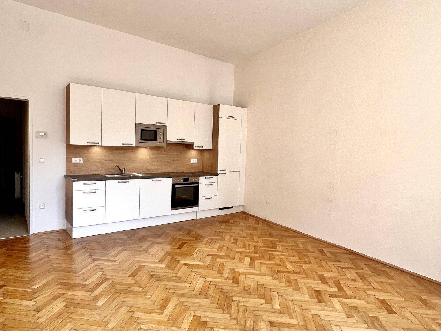 Kompakte Einbauküche mit weißen Schränken und Holzrückwand in einem Raum mit Fischgrätparkett.