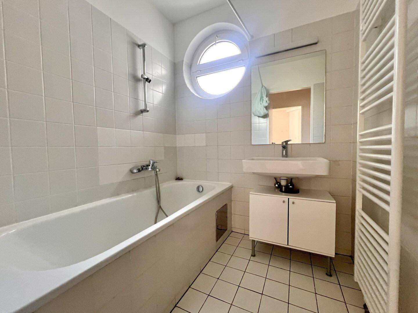 Badezimmer mit Badewanne, Waschbecken, runden Fenster und weiß gefliesten Wänden.