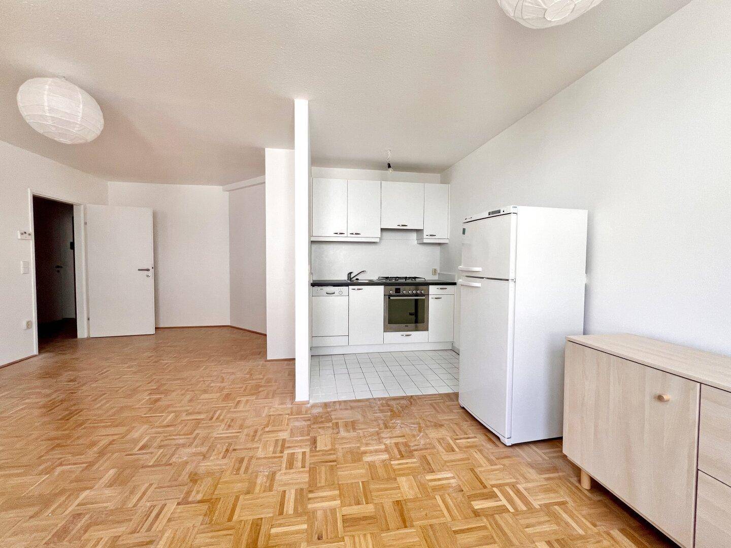 Offener Wohnbereich mit integrierter weißer Küche, ausgestattet mit Herd und Kühlschrank, auf Parkettboden.
