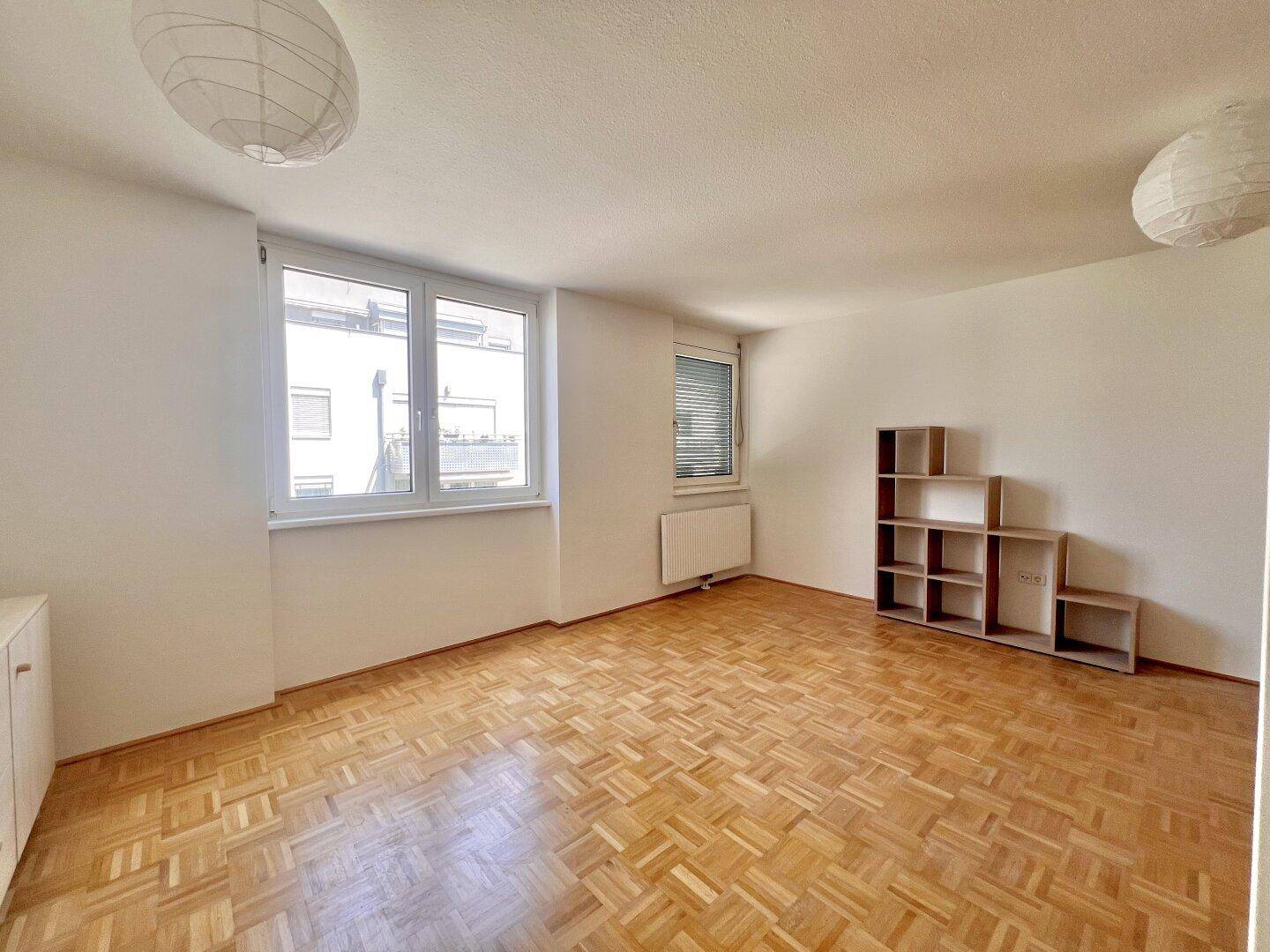 Geräumiges Wohnzimmer mit hellem Parkettboden und großen Fenstern, die viel Tageslicht hereinlassen.