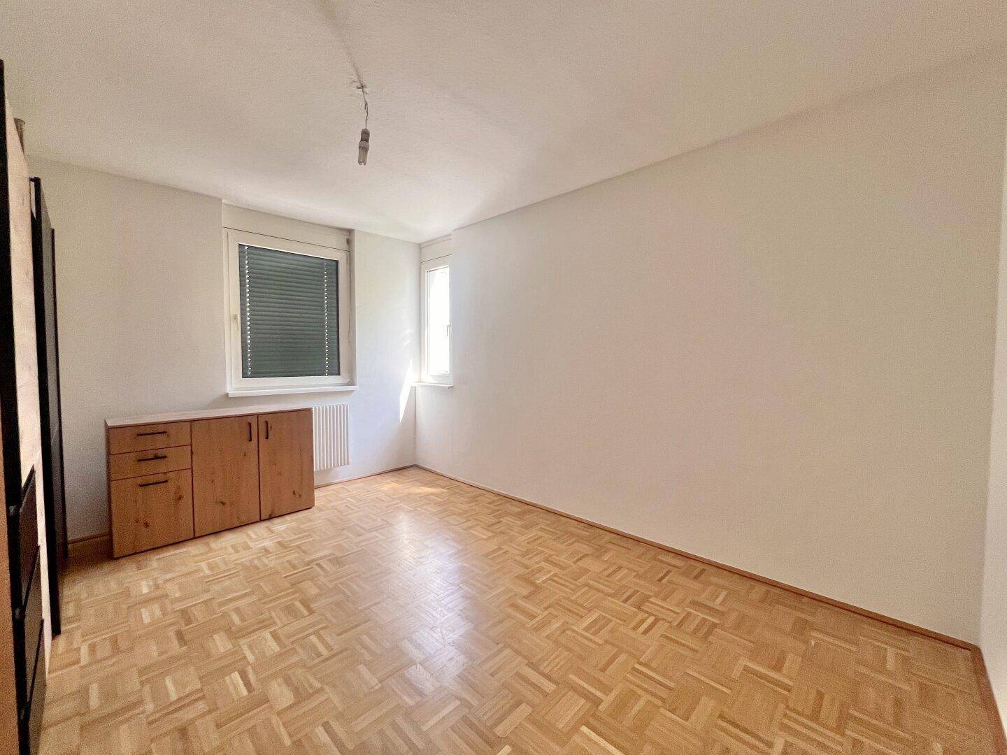 Helles Schlafzimmer mit Parkettboden und zwei Fenstern, die für eine angenehme Belichtung sorgen.