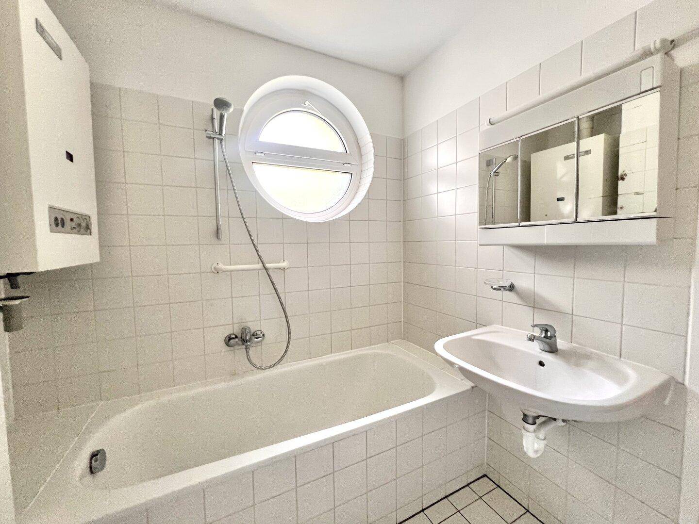Helles Badezimmer mit weißen Fliesen, Badewanne, Waschbecken und einem runden Fenster.