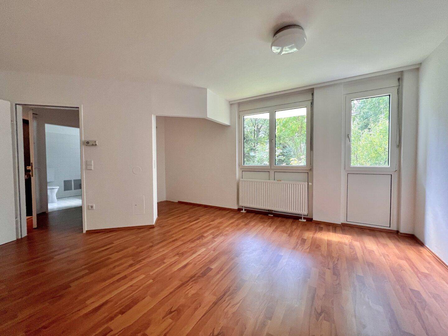 Heller Raum mit Holzboden, weißen Wänden und Blick ins Grüne durch die Fenster.