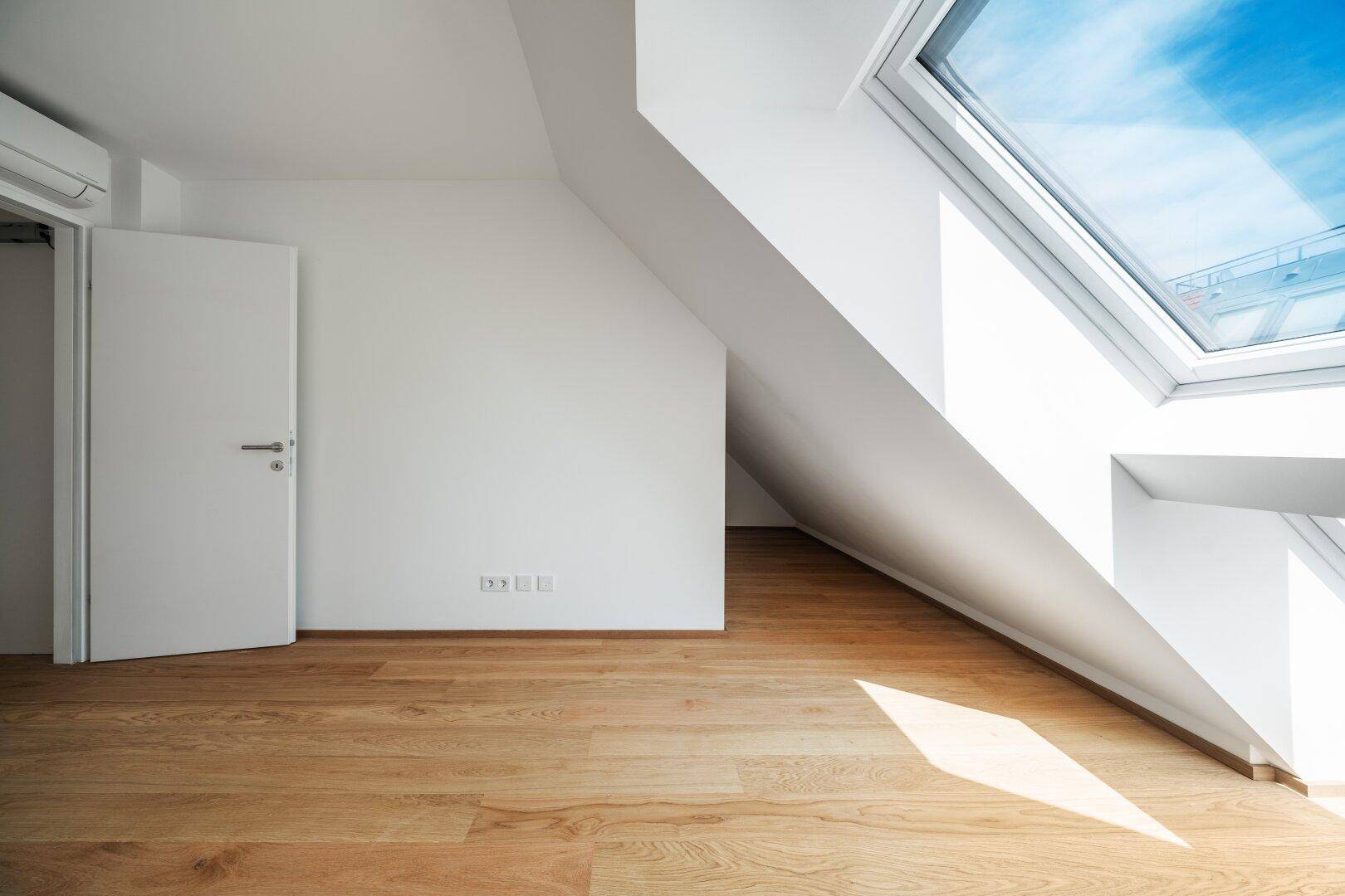 Heller Raum im Dachgeschoss mit schrägen Wänden und großem Dachfenster, bietet viel Tageslicht.