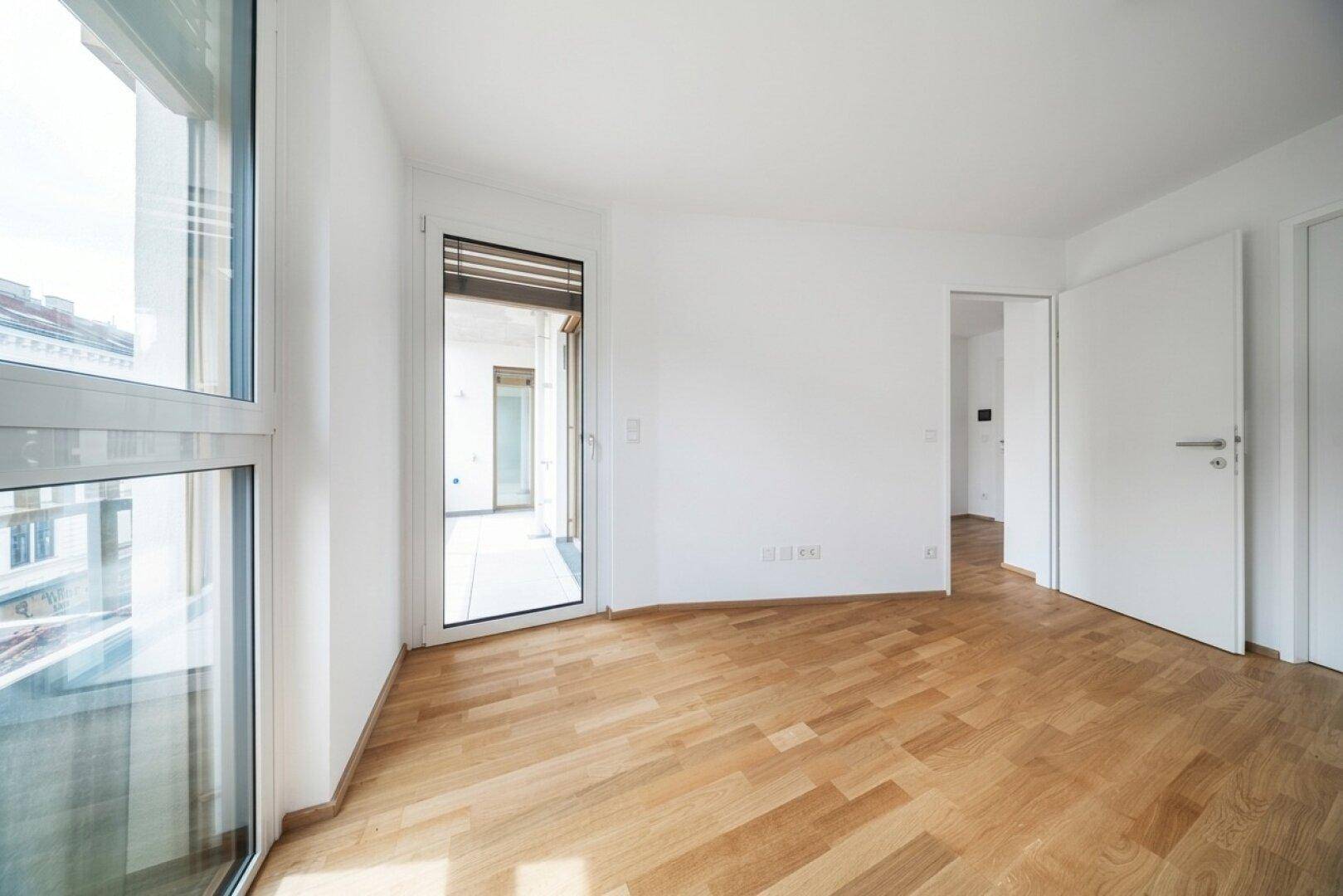 Geräumiger, heller Raum mit Parkettboden und großen Fenstern, die viel Licht hereinlassen.