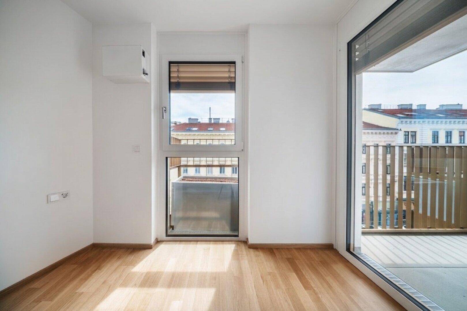 Heller Raum mit Parkettboden und Zugang zu einem Balkon, mit Blick auf die Stadt.