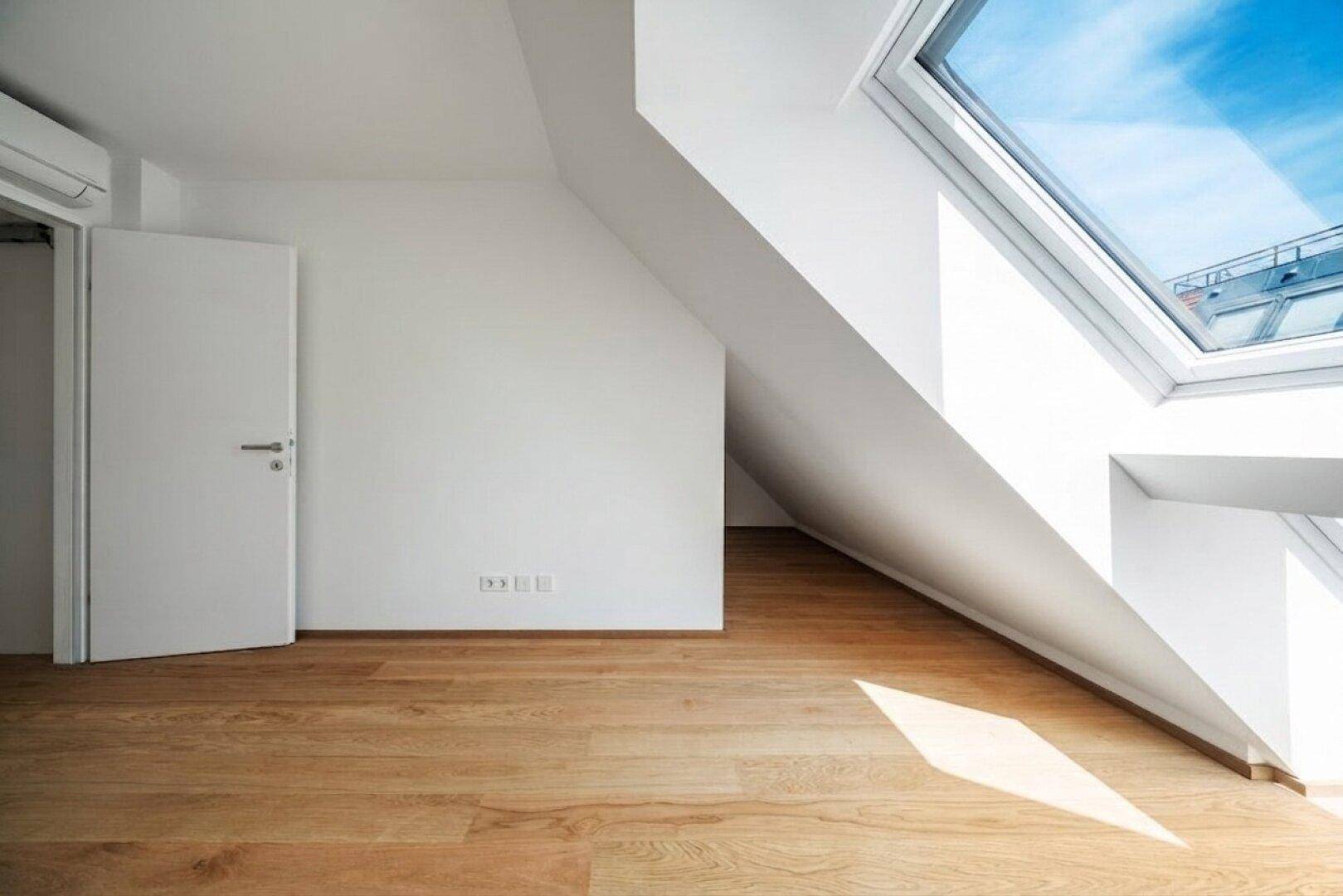 Dachzimmer mit Holzboden, schrägen Wänden und einem großen Dachfenster, das für viel Helligkeit sorgt.