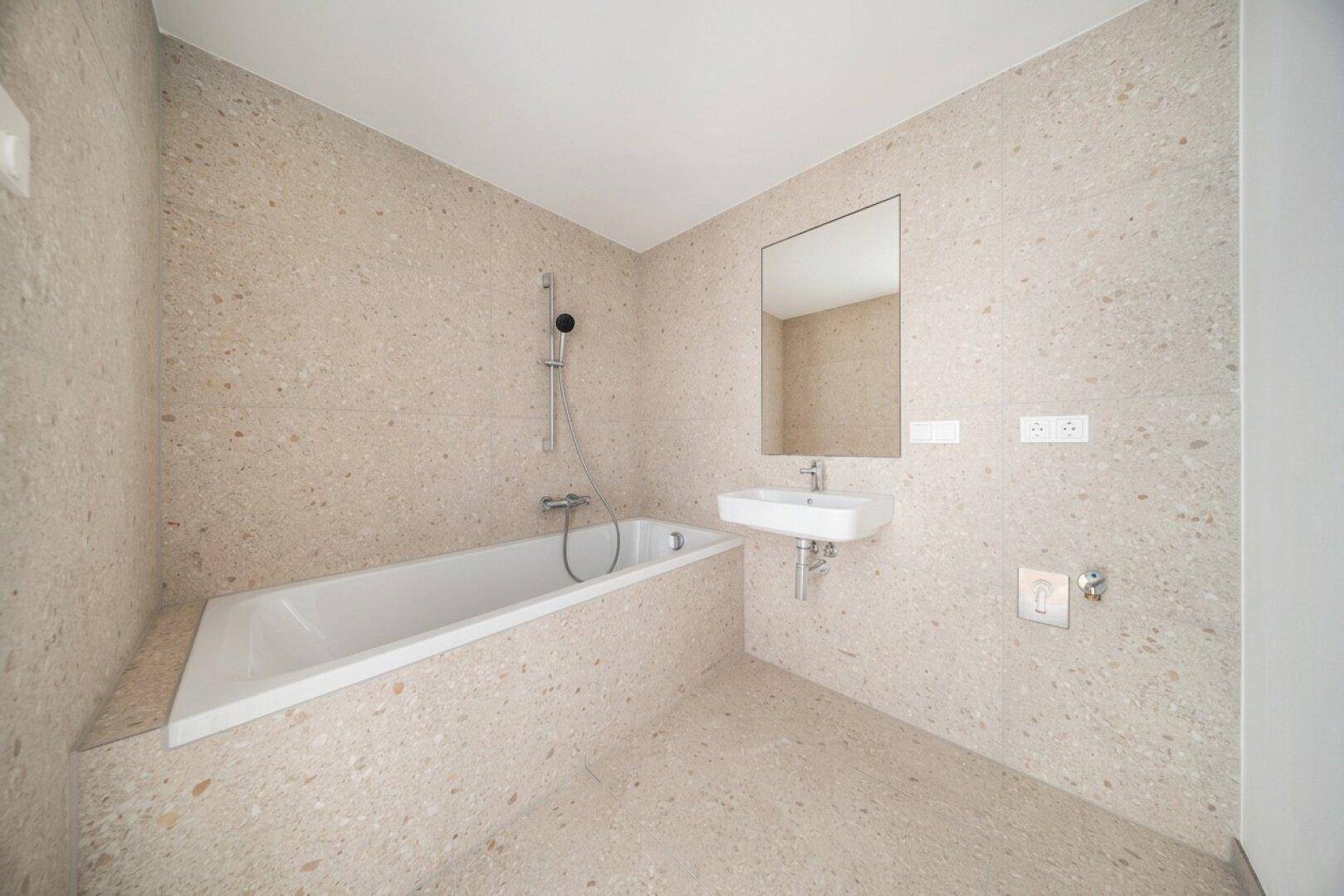 Modernes Badezimmer mit hellbeigen Fliesen, einer Badewanne und einem wandmontierten Waschbecken mit Spiegel.