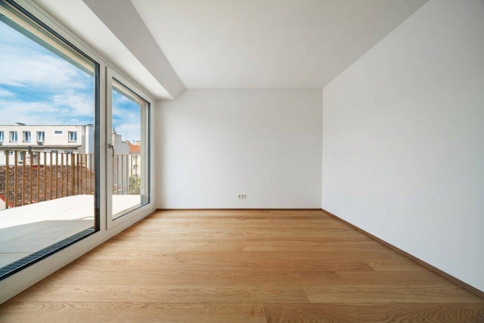 Leeres Zimmer mit Holzboden und großen Fenstern, die einen Blick auf die umliegenden Gebäude und den Himmel bieten.