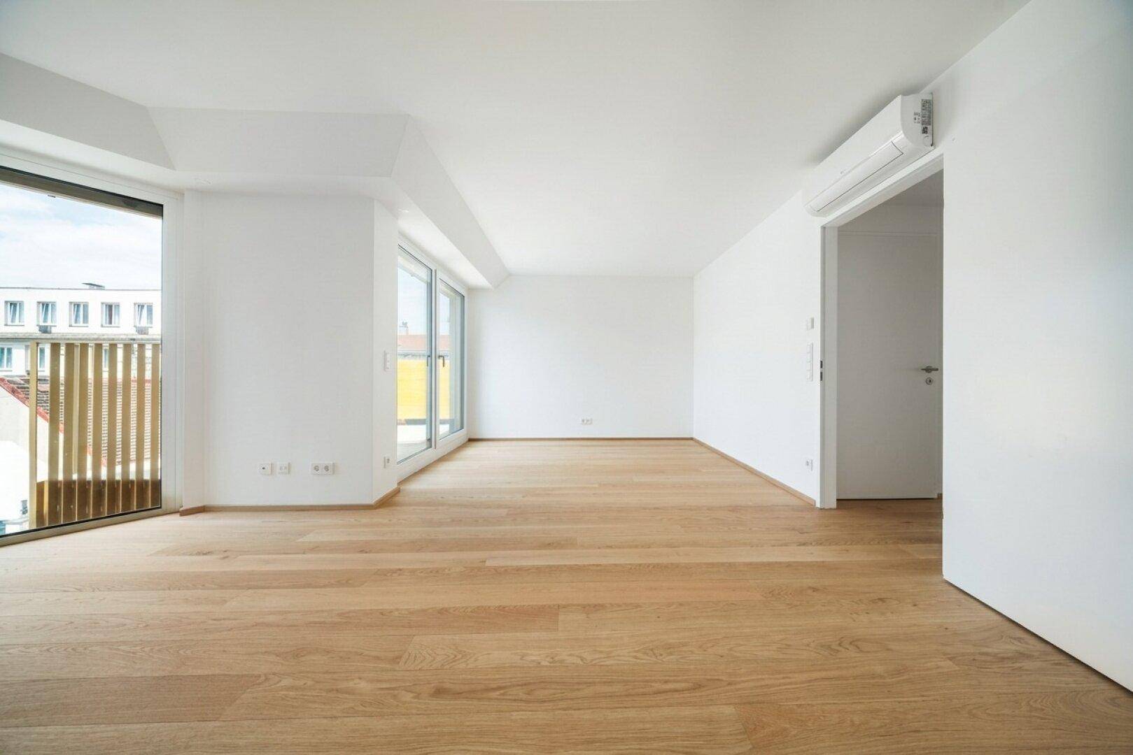 Geräumiges, helles Zimmer mit Holzboden und großen Fenstern, die viel Tageslicht hereinlassen und Zugang zum Außenbereich bieten.
