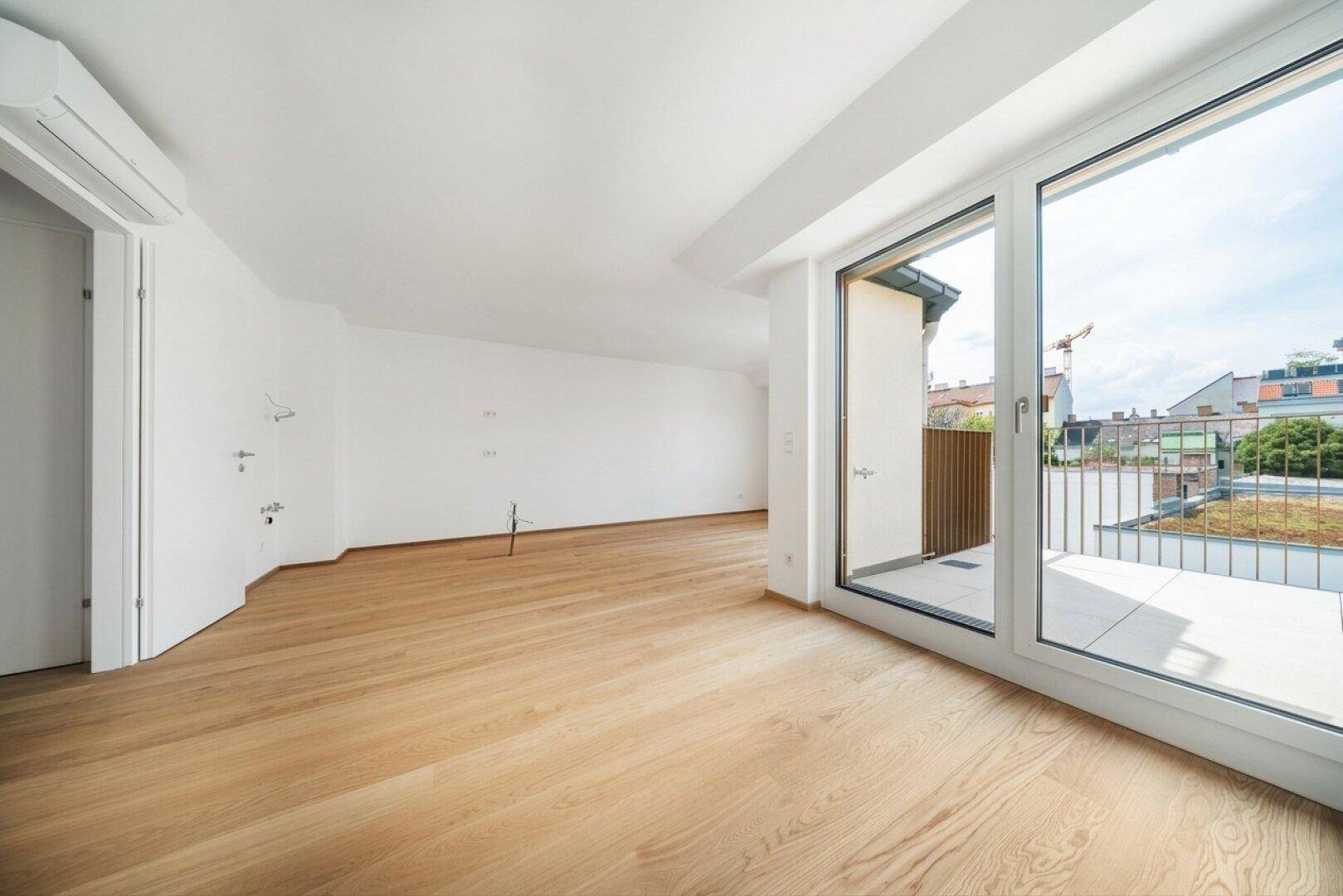 Geräumiges, helles Zimmer mit Holzboden und großen Fenstern, die viel Tageslicht hereinlassen und Zugang zum Außenbereich bieten.