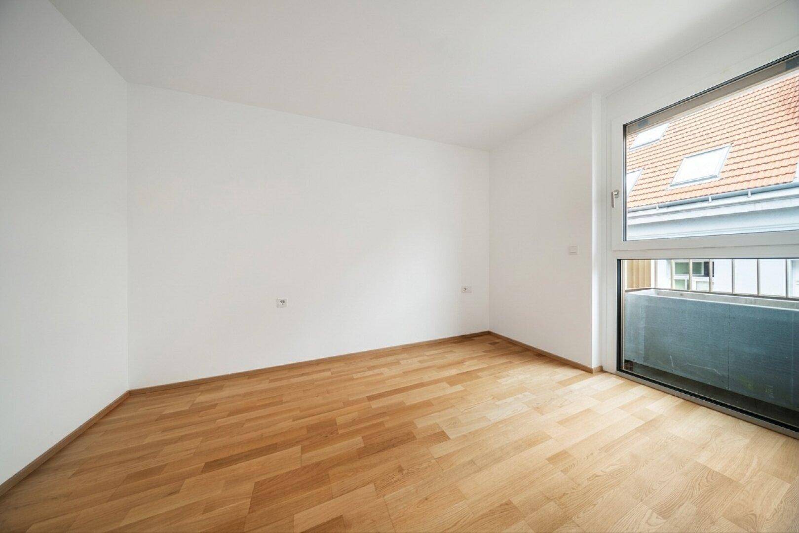 Leeres Schlafzimmer mit Parkettboden und großem Fenster, das viel Tageslicht hereinlässt.