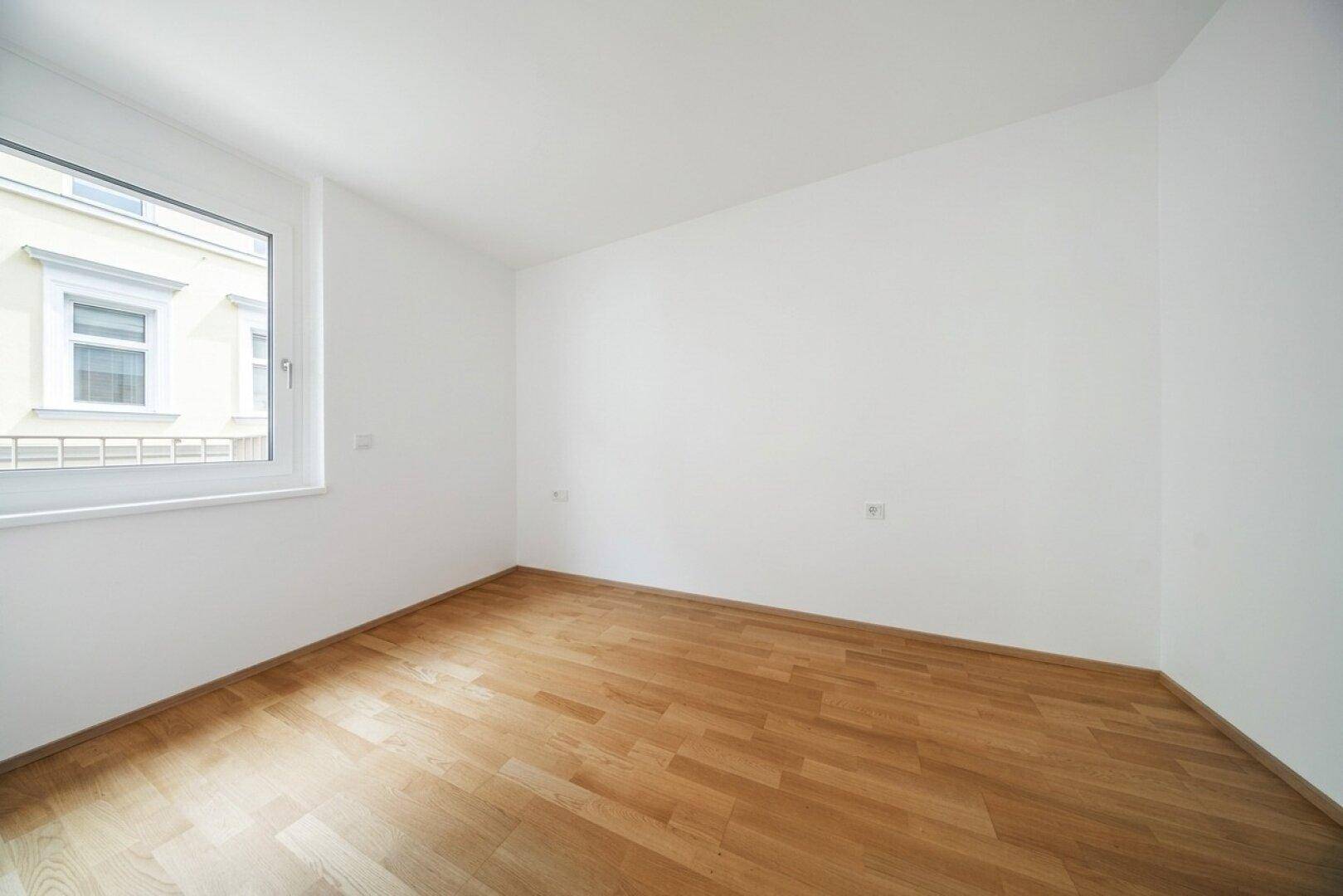 Unmöbliertes Zimmer mit Parkettboden und einem großen Fenster, das viel Licht spendet.