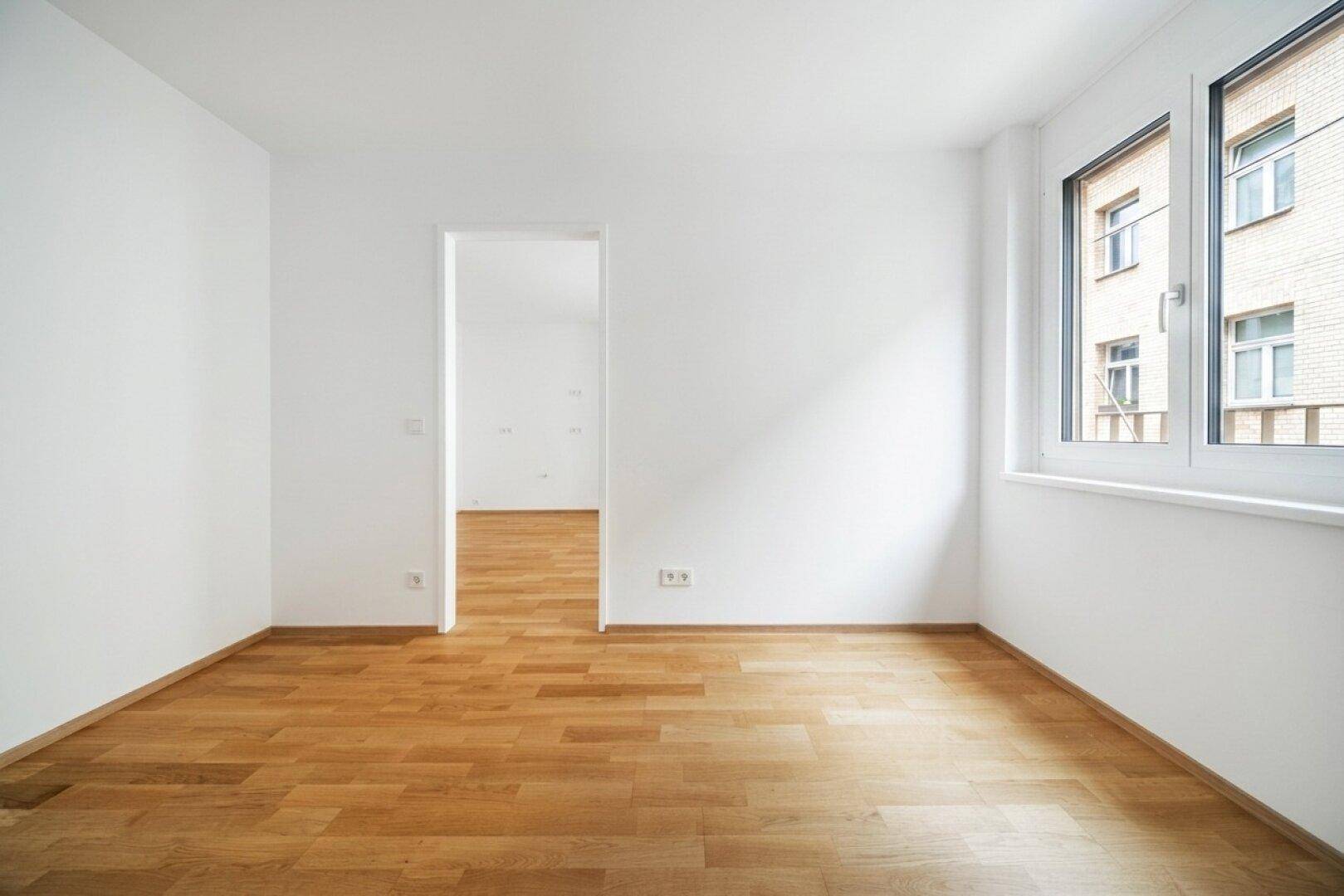 Leerer Raum mit hellem Parkettboden und weiß gestrichenen Wänden, mit Zugang zu einem weiteren Zimmer.