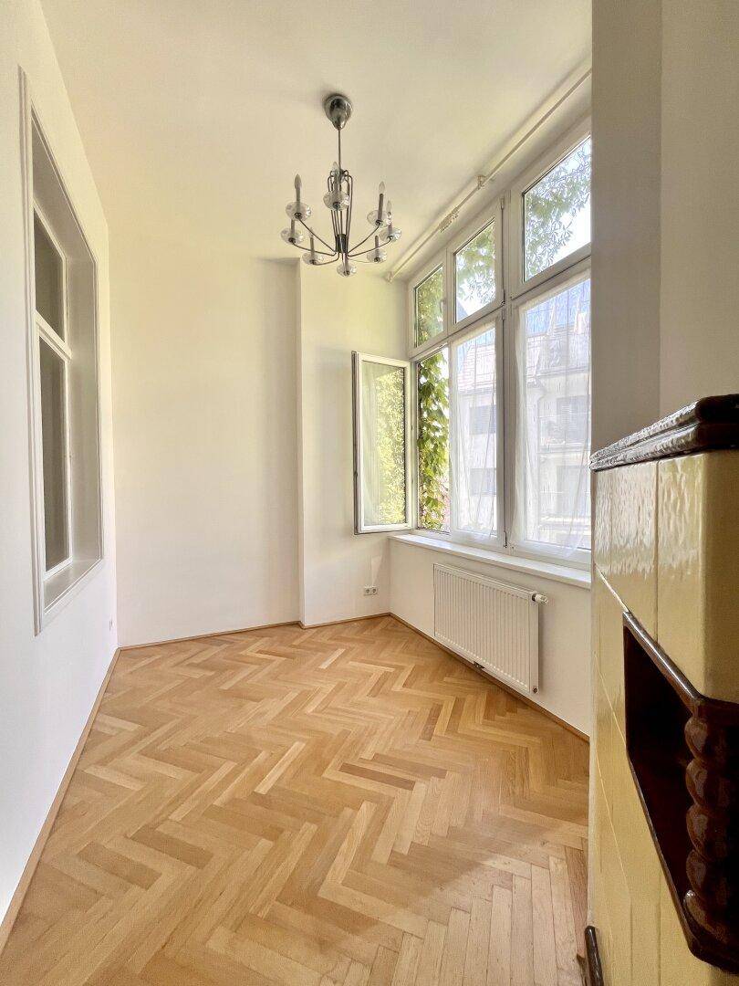 Helles Zimmer mit Fischgrätparkett, Kachelofen und großen Fenstern mit Blick ins Grüne.