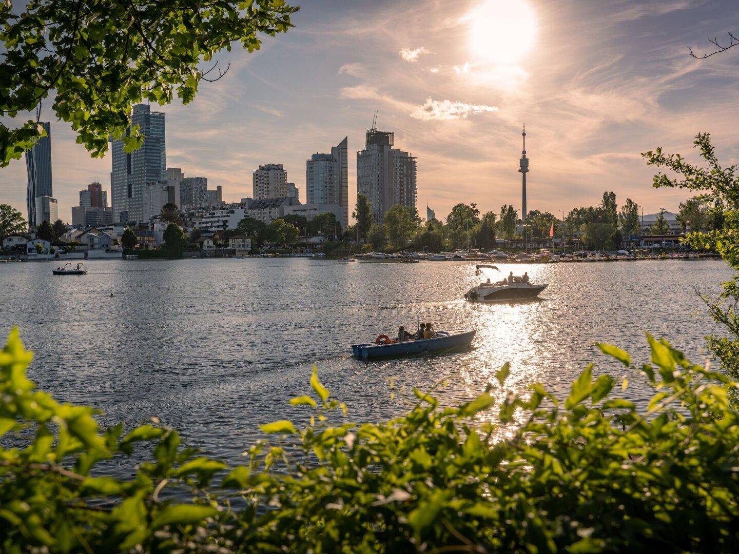 Städtische Skyline am Wasser mit Booten bei Sonnenuntergang, umrahmt von grünen Bäumen und hellem Licht.