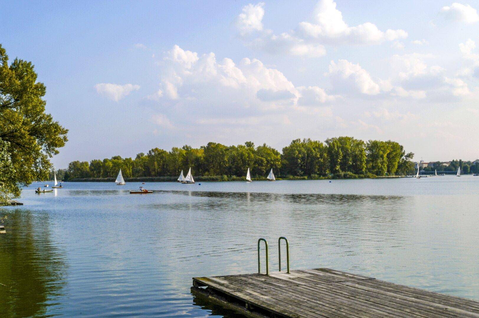 Ruhiger See mit Segelbooten und bewaldeter Insel unter blauem Himmel, mit Holzsteg im Vordergrund.