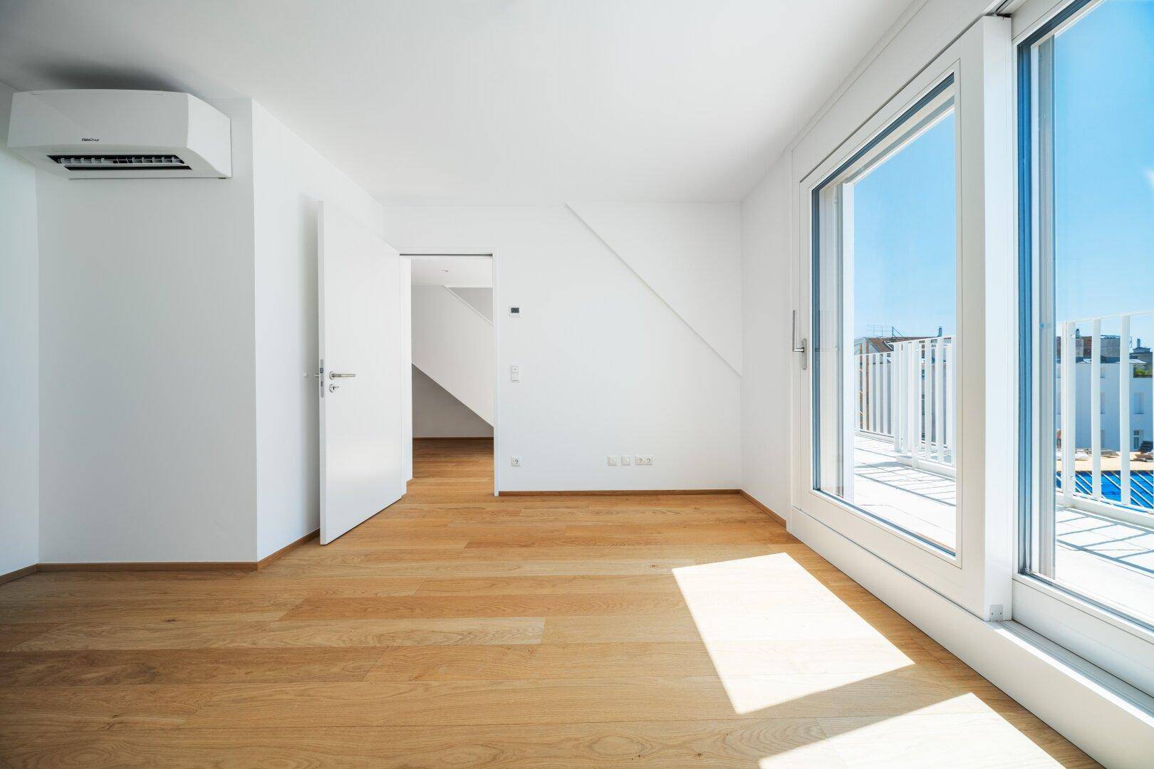 Lichtdurchfluteter Raum mit Holzboden, Zugang zum Balkon und Klimaanlage.